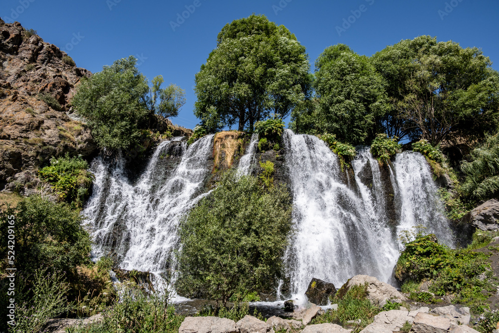 a stormy waterfall against the backdrop of green trees and mountains and a blue sky in the mountains of Armenia