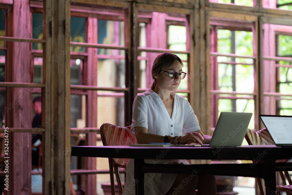 Focused concentrated female freelancer wearing eyeglasses sitting at desk in coworking space, looking at laptop screen, concentrated businesswoman in dress working on computer in open plan office Focused concentrated female freelancer wearing eyeglasses sitting at desk in coworking space, looking at laptop screen, concentrated businesswoman in dress working on computer in open plan office