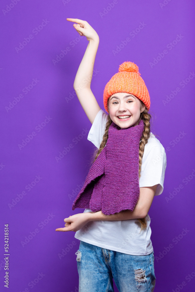 Winsome Caucasian Teenager Girl in Coral Warm Knitted Winter Hat and Violet Scarf Showing Direction With Both Hands Over Purple Background. Winsome Caucasian Teenager Girl in Coral Warm Knitted Winter Hat and Violet Scarf Showing Direction With Both Hands Over Purple Background.