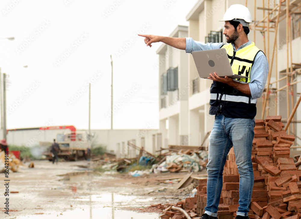 Bearded man engineer wears safety helmet inspecting housing development project at construction site using laptop computer. Contractor manager examining residential building estate infrastructure. Bearded man engineer wears safety helmet inspecting housing development project at construction site using laptop computer. Contractor manager examining residential building estate infrastructure.