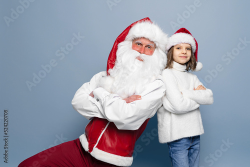 Cute little girl in Christmas hat with Santa looking at camera against blue studio background. Isolate