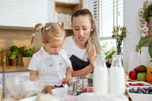 Preschooler daughter with young mom wearing aprons decorating cupcakes with berries.Girls cooking baking homemades muffing in modern beautiful light kitchen using eggs milk berries.