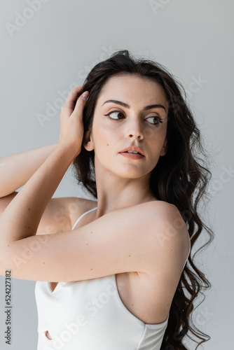 Young woman touching brunette hair and looking away isolated on grey.
