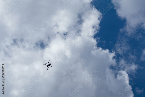 Drone quadrocopter silhouette flying high in white clouds on blue sky, cloudscape background. Skyscape scenery