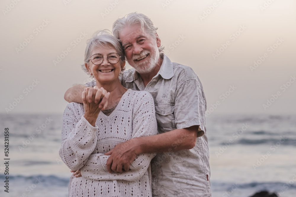 Portrait of two beautiful and happy seniors or retirees embraced on the beach in sunset light - old smiling senior couple outdoors enjoying holidays together