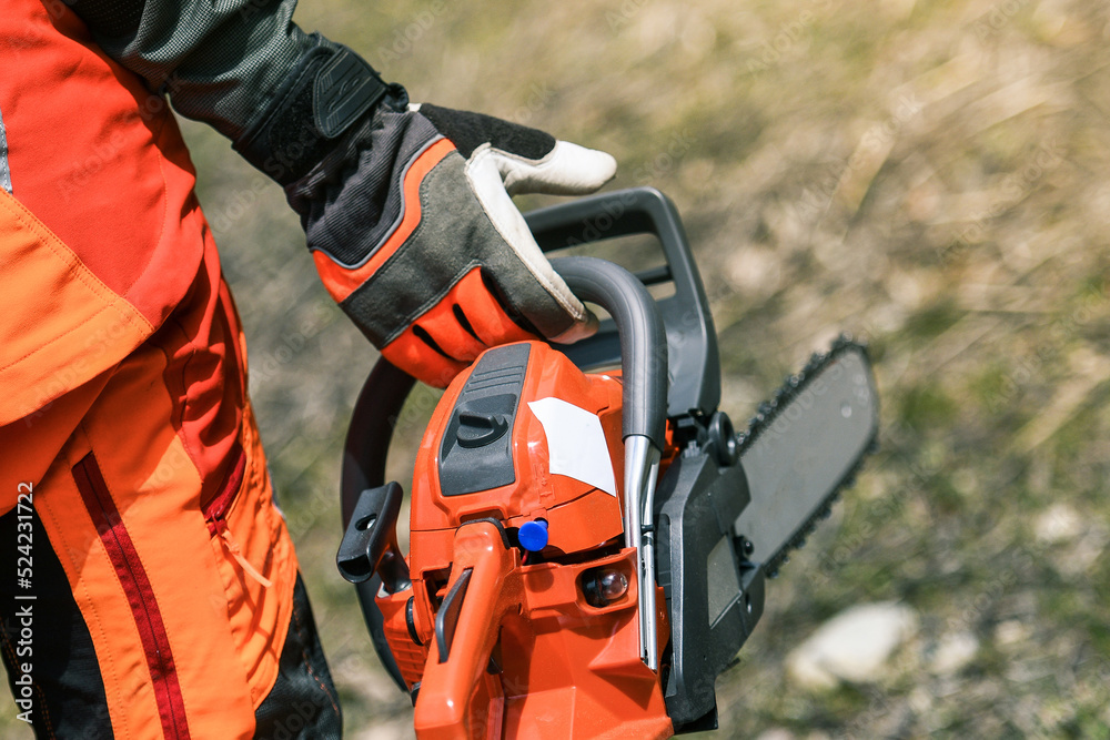 Close up view of an hand holding a chainsaw. Lumberjack at work wears orange personal protective equipment. Gardener working outdoor in the forest. Security, professionalism, occupation worker concept Close up view of an hand holding a chainsaw. Lumberjack at work wears orange personal protective equipment. Gardener working outdoor in the forest. Security, professionalism, occupation worker concept