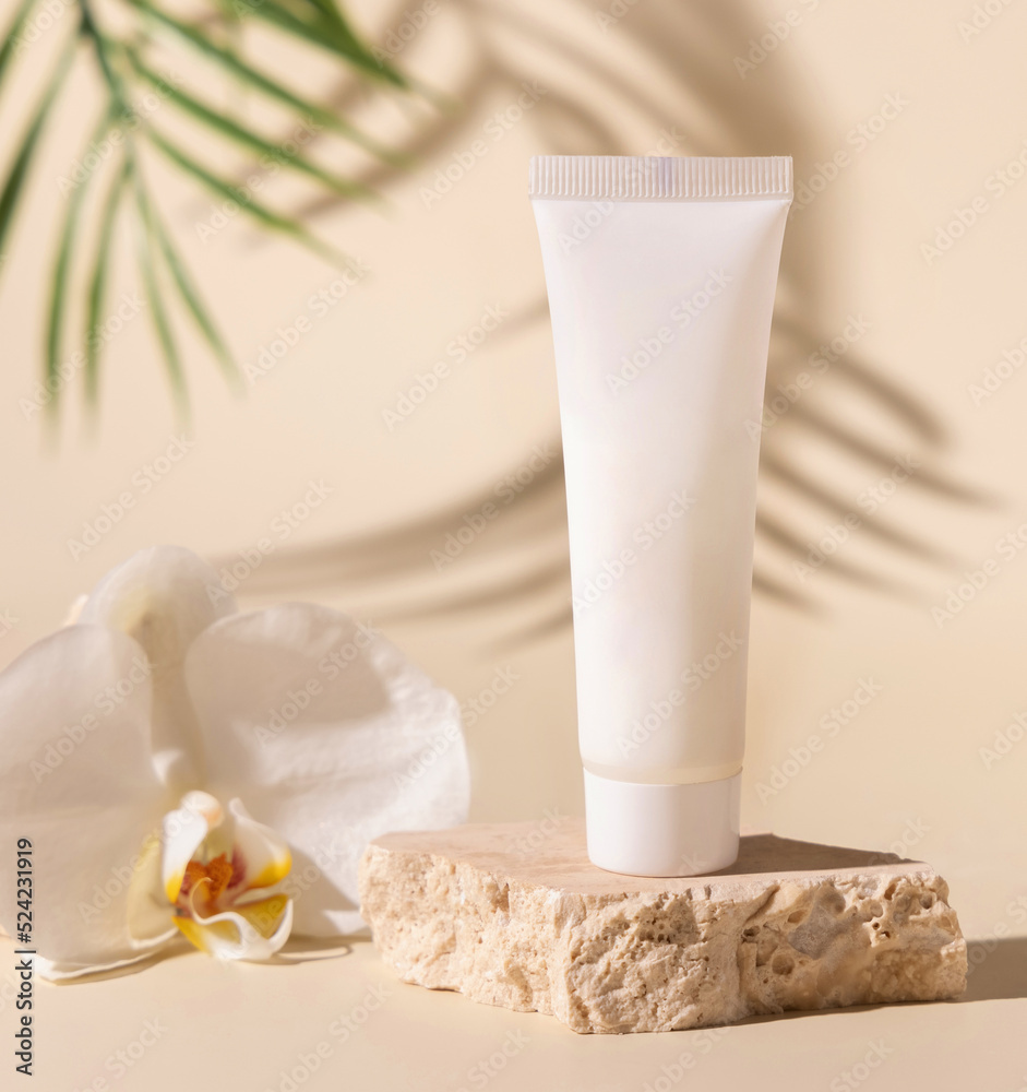 White plastic cream tube on stone near orchid flower on light beige, hard shadows. Mockup White plastic cream tube on stone near orchid flower on light beige, hard shadows. Mockup