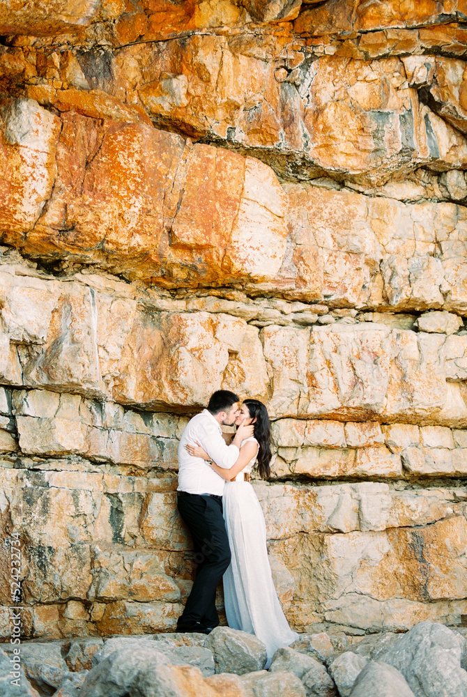 Groom kisses bride holding her face with his hands near a huge stone wall
