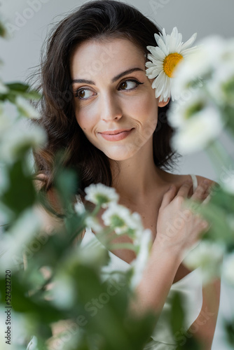 Fotografia Smiling woman with visage and chamomile in hair looking away near blurred plants isolated on grey