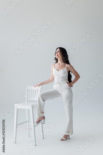 Smiling woman in white clothes posing near chair on grey background.