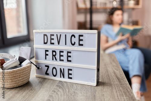 digital detox and leisure concept - close up of device free zone words on light box, gadgets in basket on table and woman reading book at home