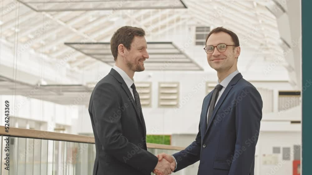 Tilt up medium slowmo portrait of two Caucasian mid aged businessmen in formalwear shaking hands and smiling at camera standing by glass railing at modern business center Tilt up medium slowmo portrait of two Caucasian mid aged businessmen in formalwear shaking hands and smiling at camera standing by glass railing at modern business center