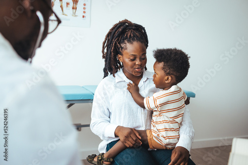 Pediatrician's Office. Shot of a single mother bringing his little boy for a checkup. An adorable toddler boy sits in his mother's lap in his pediatrician's office. He smiles and looks away. 