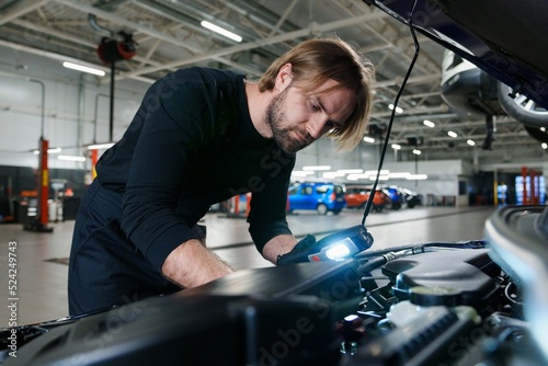 A male mechanic in uniform with a flashlight performs diagnostics under the hood of the car. Modern and technological car service