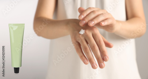 Woman applying cream and tube of hand care cosmetic product on light background, closeup