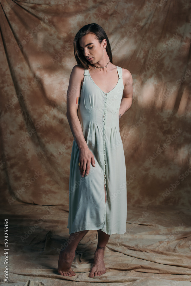 Barefoot queer person in sundress standing on abstract brown background.