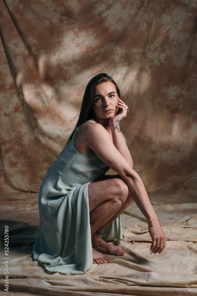 Trendy queer person in sundress looking at camera while posing on abstract brown background. Trendy queer person in sundress looking at camera while posing on abstract brown background.