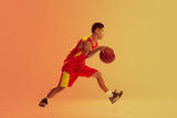 Dynamic portrait of young man, basketball player in motion, dribbling isolated over orange studio background in neon light