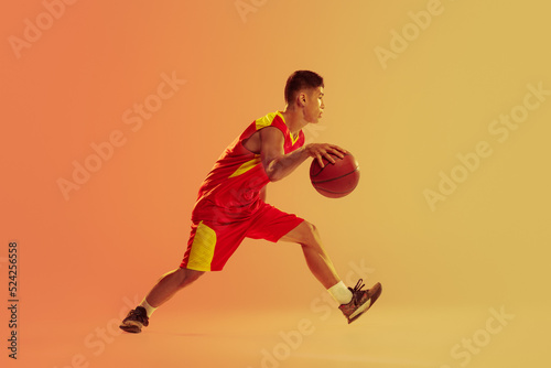 Dynamic portrait of young man, basketball player in motion, dribbling isolated over orange studio background in neon light