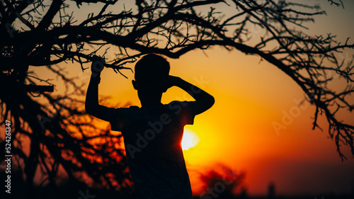The silhouette of a man looking into the distance from under the palm of his hand against the background of the sunset sky under the branches of a tree