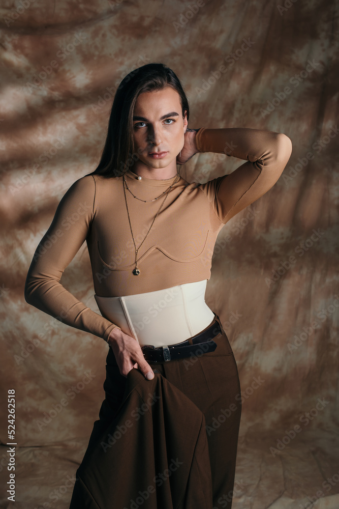 Trendy nonbinary person holding blazer and posing on abstract brown background. Trendy nonbinary person holding blazer and posing on abstract brown background.