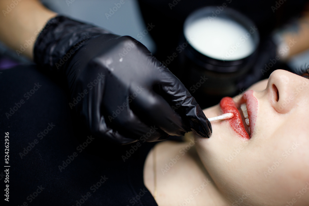 Cosmetologist applying vaseline petrolatum to permanent make up tattoo on young woman lips. Permanent lips tattoo procedure concept