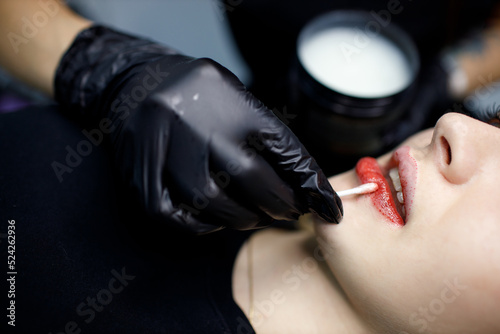 Cosmetologist applying vaseline petrolatum to permanent make up tattoo on young woman lips. Permanent lips tattoo procedure concept photo