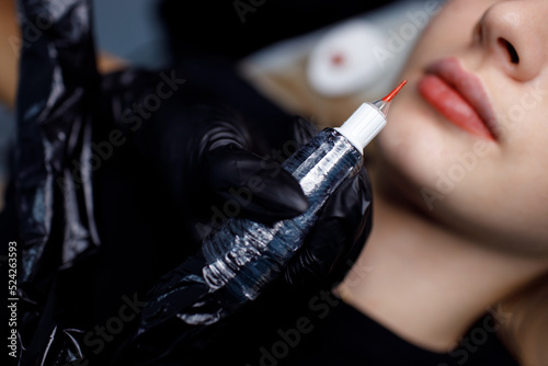 Cosmetologist applying red permanent make up tattoo on young woman lips.