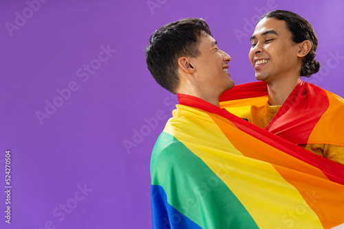 Happy biracial male couple embracing and holding lgbt flag on purple background