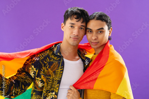 Portrait of happy biracial male couple embracing and holding lgbt flag on purple background