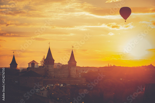 Bright orange sunset over Kamianets-Podilskyi castle fortress. One hot air balloon silhouette against golden sky, watching evening old town cityscape. Travel background. Popular touristic destination photo
