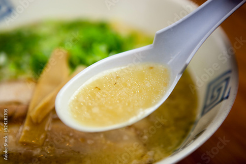 ラーメン店で提供される、背脂が浮いたコク深い黄金色の塩ラーメンスープ。レンゲですくい上げたシズル感あふれるアップ写真