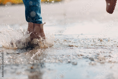 Woman wearing rain rubber boots walking running and jumping into puddle with water splash and drops in autumn rain.