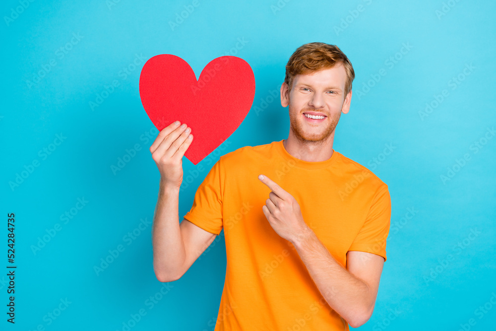 Photo of nice cute boyfriend man toothy smile arm direct index present large red paper love symbol sign isolated on cyan color background Photo of nice cute boyfriend man toothy smile arm direct index present large red paper love symbol sign isolated on cyan color background