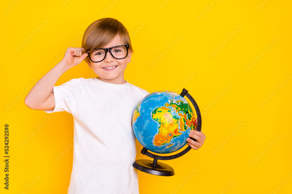 Portrait of handsome cheerful diligent pre-teen boy holding globe touching specs isolated over bright yellow color background Portrait of handsome cheerful diligent pre-teen boy holding globe touching specs isolated over bright yellow color background