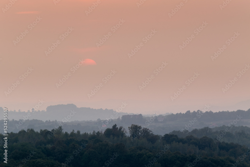 Atmospheric landscape scene of lush forest layers in mist during autumn sunset in countryside, forest obscured in fog and big orange sun dimmed behind clouds