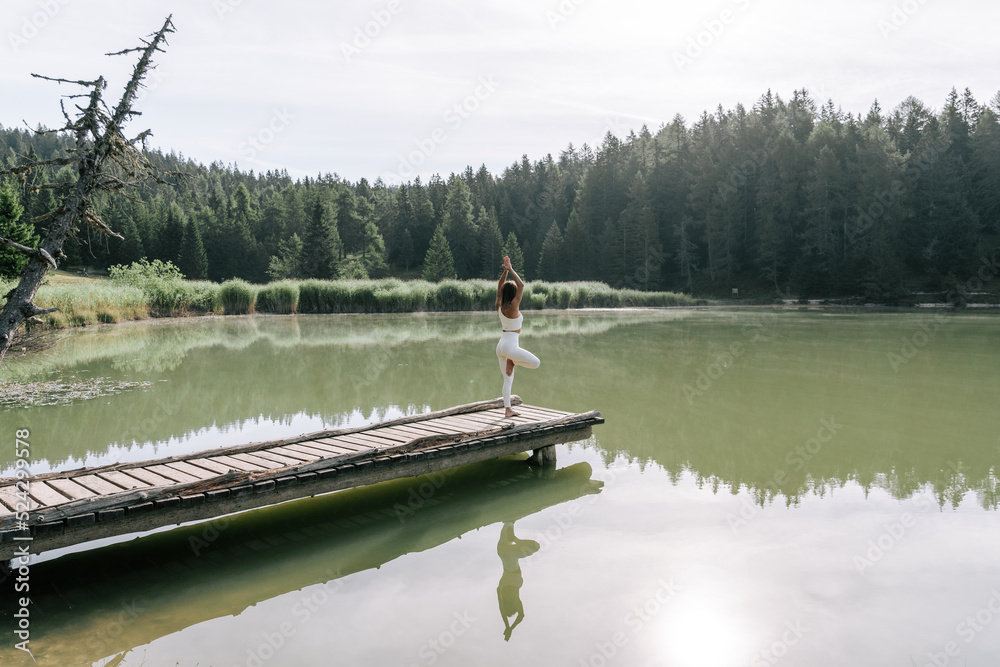 Eine Junge Frau praktiziert Yoga auf einem Steg am See zum Sonnenaufgang. Der Felixer Weiher - Tretsee in Südtirol. Weiße Leggins 5
