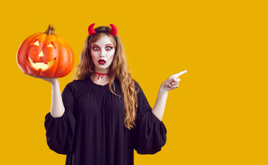 Portrait young woman in Halloween costume standing isolated on yellow background, holding Jack o lantern, pointing finger at copyspace side and looking at camera with funny surprised face expression