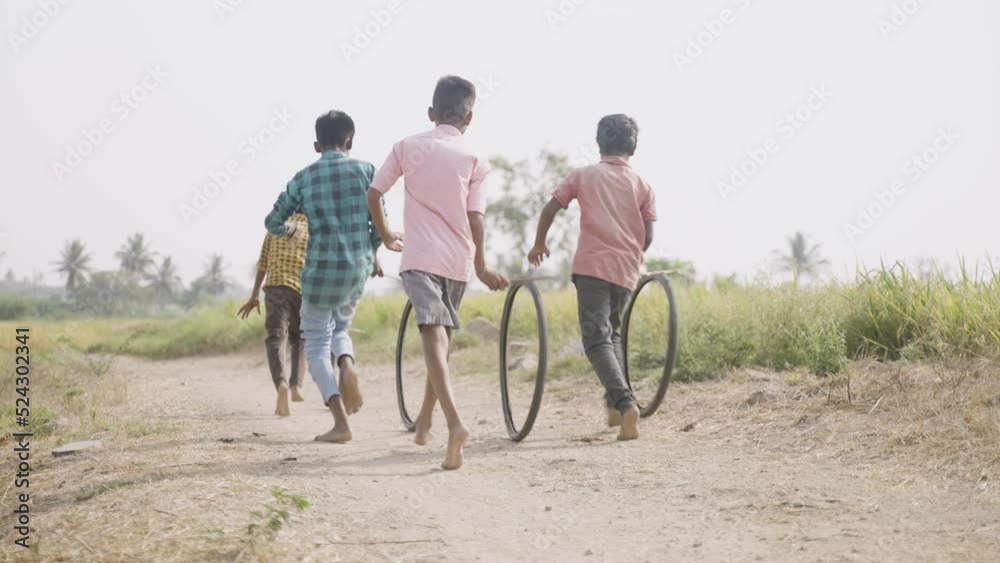 back view shot of Indian village kids playing with tyre wheel by rolling near paddy agricultural field - concept of playful village lifestyle, outdoor activities and rural india. back view shot of Indian village kids playing with tyre wheel by rolling near paddy agricultural field - concept of playful village lifestyle, outdoor activities and rural india.