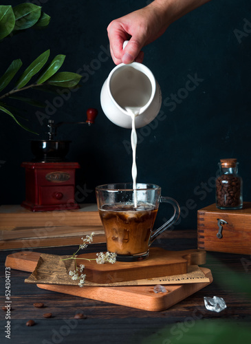 Mixing cold black coffee with milk in a glass mug. Add milk from a milk jug to coffee. Dark key, low key. copy space. Blue background. Coffee grains on the table