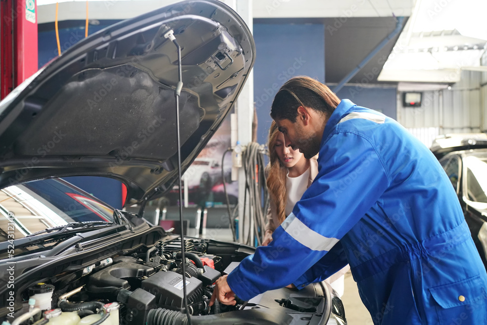 Male car operator wearing blue overalls,cap and gloves working under the hood of white car and checking attentively serviceability of engine at repair garage. Concept of car maintenance Male car operator wearing blue overalls,cap and gloves working under the hood of white car and checking attentively serviceability of engine at repair garage. Concept of car maintenance