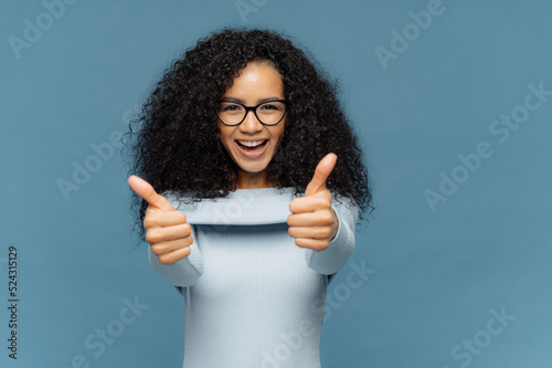 Happy lovely woman with bushy Afro hair gives thumb up, approves nice idea, wears fashionable sweater, isolated over blue background. Its fine, I agree. Body language concept. People and like concept © VK Studio