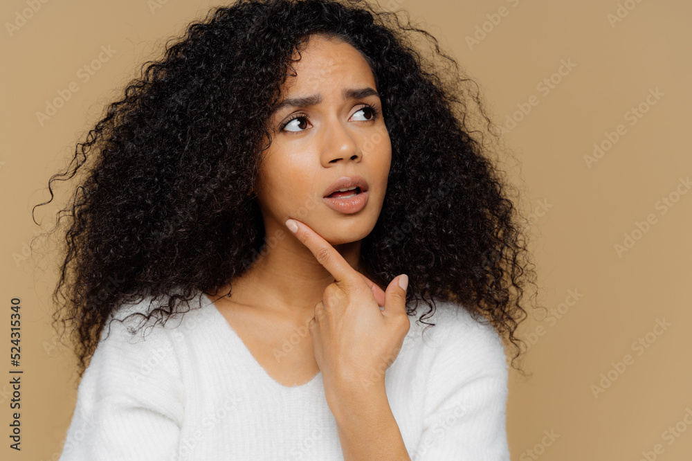 Puzzled female model keeps fore finger on chin, looks thoughtfully aside, has frustrated facial expression, curly Afro hair, wears casual white jumper, isolated over brown background. Close up shot Puzzled female model keeps fore finger on chin, looks thoughtfully aside, has frustrated facial expression, curly Afro hair, wears casual white jumper, isolated over brown background. Close up shot