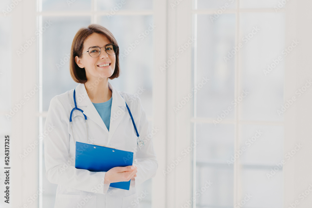 Experienced woman pediatrician stands with clipboard in cabinet, wears white medical coat with stethoscope, gives excellent medical treatment, has happy expression, ready to give consultancy Experienced woman pediatrician stands with clipboard in cabinet, wears white medical coat with stethoscope, gives excellent medical treatment, has happy expression, ready to give consultancy