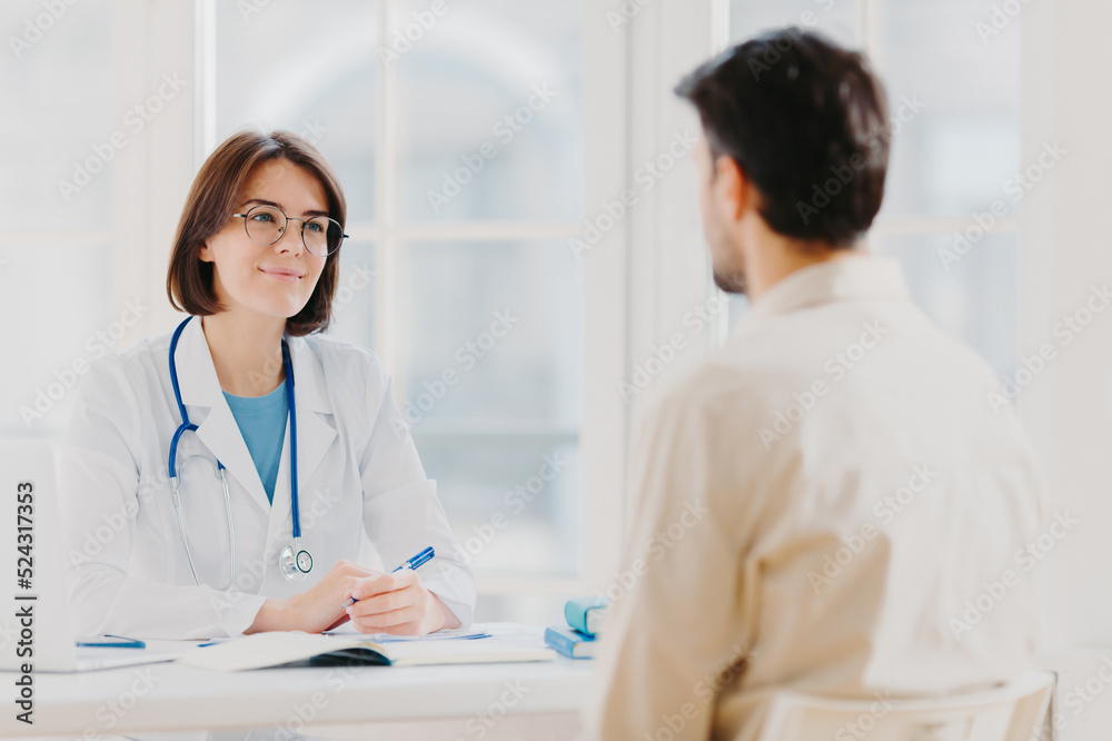 Doctor and patient discuss something, sit at table in clinic. Female cardiologist in eyewear gives medical consultation diagnostic, advice for man how to cure disease, pose in hospital room. Doctor and patient discuss something, sit at table in clinic. Female cardiologist in eyewear gives medical consultation diagnostic, advice for man how to cure disease, pose in hospital room.