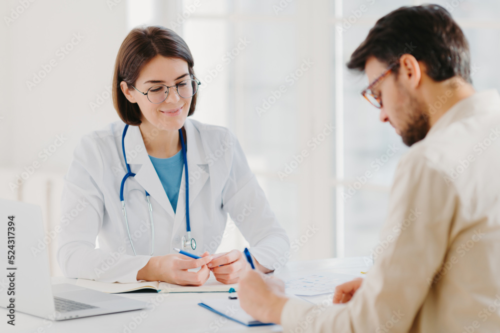 Male patient writes down application form, gets consultancy and advice about home treatment from doctor, use laptop and medical document at desk, pose at office together. Man doctors appointment Male patient writes down application form, gets consultancy and advice about home treatment from doctor, use laptop and medical document at desk, pose at office together. Man doctors appointment