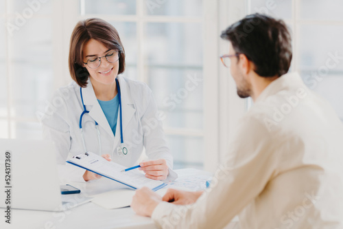 Medical appointment. Female doctor gives professional medical help to male patient, explains written information on paper in clipboard, gives support and good service, pose at hospital near desktop. © VK Studio