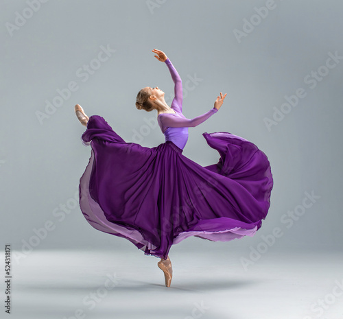 Fototapete Ballerina in purple ballet leotard and violet long ballet skirt dancing in white studio on pointe shoes