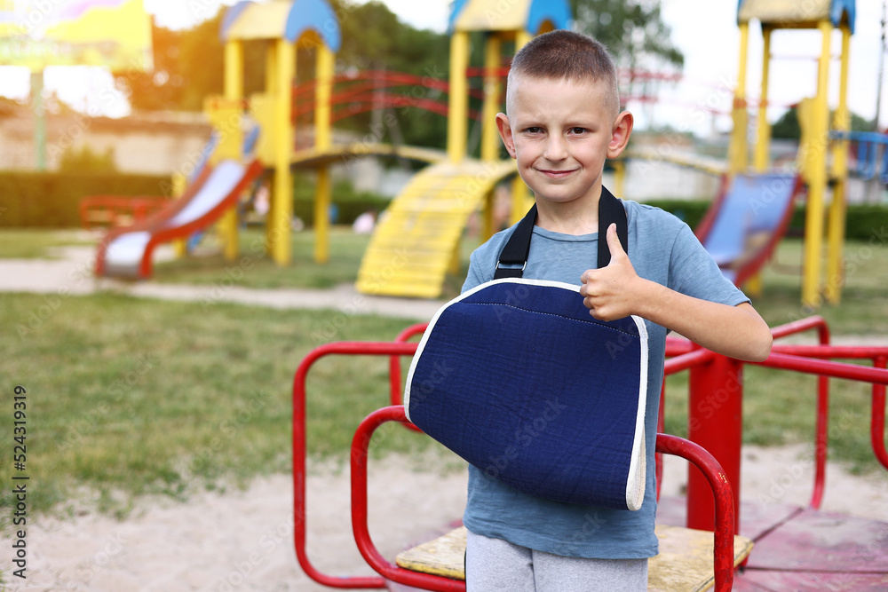 Fototapeta premium happy child with broken limb arm outdoors on playground background. school boy had accident on summer vacation. kid in a cast on hand. concept of health and medical.