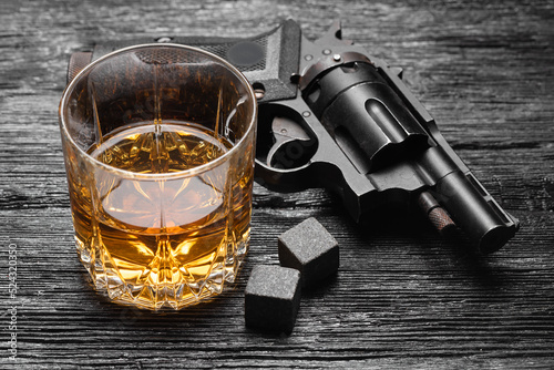 Glass of whiskey and black gun on the wooden table background.